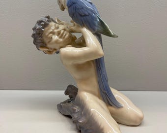 May include: A porcelain figurine of a kneeling boy with a blue and white parrot perched on his shoulder. The boy has a mischievous expression on his face and the parrot is looking at him.