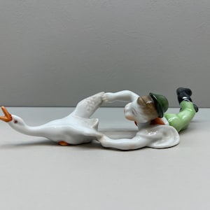 Puede incluir: Una figurita de porcelana de un niño con un sombrero verde y pantalones marrones luchando con un ganso blanco.