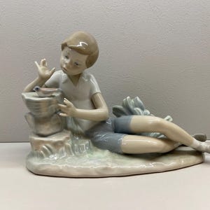 Può includere: Statuina in porcellana raffigurante un giovane reclinato, che interagisce con un uccello. La figura indossa una camicia bianca e pantaloncini grigi. L'uccello è posato su una struttura in pietra. Finitura opaca.