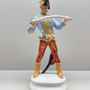 Puede incluir: Una figurita de porcelana de un soldado con un uniforme azul y dorado, sosteniendo una espada. Lleva un sombrero rojo y dorado y pantalones rojos con ribetes dorados. La figurita está de pie sobre una base blanca.