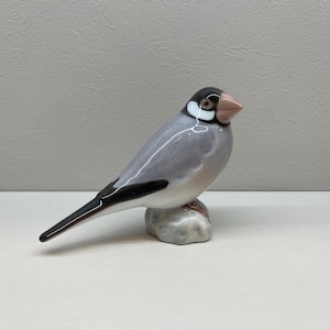 Peut inclure: Une figurine en céramique représentant un moineau de Java perché sur un socle en forme de rocher. L'oiseau présente un corps gris lisse, une tête noire avec des taches blanches sur les joues, un bec rose pâle et une longue queue noire.
