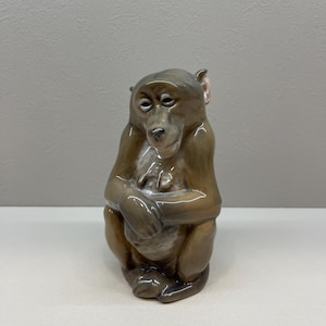 Puede incluir: Figura de cerámica de un mono en posición sentada. El mono es marrón con un acabado brillante y los brazos cruzados. La figura tiene rasgos faciales detallados y una expresión pensativa. El fondo es gris claro liso.