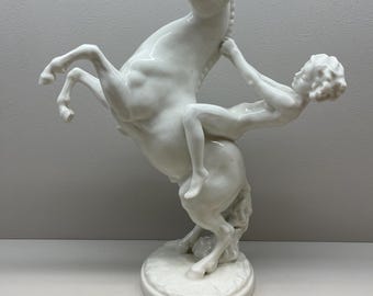 May include: A white porcelain sculpture depicting a rearing horse and a figure. The horse is on its hind legs, with the figure reaching for the horse's head. The sculpture is mounted on a round base. A decorative item.