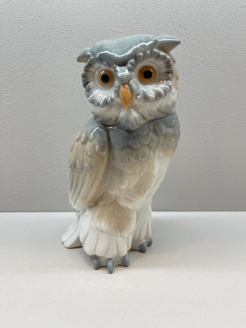 K&ouml;nnte beinhalten: Eine Eulenfigur aus Porzellan mit gl&auml;nzender Oberfl&auml;che in den Farben sanftes Grau, Wei&szlig; und Beige. Die Eule hat gro&szlig;e, runde, bernsteinfarbene Augen und einen gelben Schnabel, mit detaillierten Federstrukturen, die &uuml;ber den K&ouml;rper geformt sind.