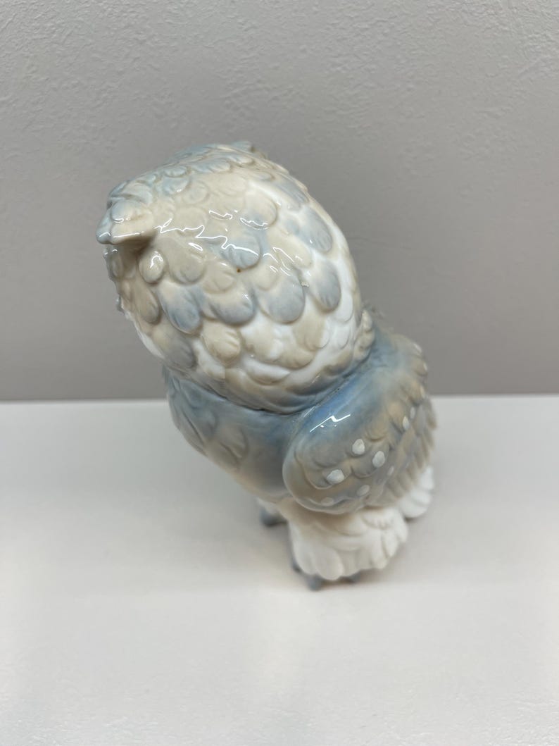 K&ouml;nnte beinhalten: Eine Eulenfigur aus Porzellan mit gl&auml;nzender Oberfl&auml;che in den Farben sanftes Grau, ged&auml;mpftes Blau und Wei&szlig;. Die Eule ist aus einer hohen R&uuml;ckansicht dargestellt und zeigt detaillierte, modellierte Federstrukturen an Kopf und Fl&uuml;geln.