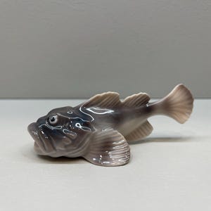 May include: A ceramic figurine of a fish with a detailed design. The fish has a gray body with lighter fins and tail. The figurine is resting on a white surface.