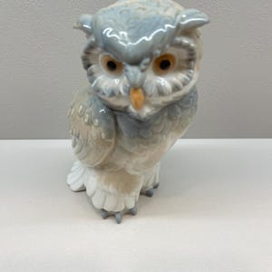 K&ouml;nnte beinhalten: Eine Eulenfigur aus Porzellan mit gl&auml;nzender Oberfl&auml;che und einem Gefieder in sanften Grau-, Wei&szlig;- und Beiget&ouml;nen. Die Eule hat ausgepr&auml;gte Federohren, gro&szlig;e bernsteinfarbene Augen und einen kleinen, spitzen gelben Schnabel in stehender Position.