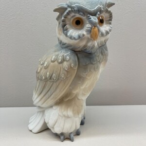 K&ouml;nnte beinhalten: Eine Eulenfigur aus Porzellan mit gl&auml;nzender Oberfl&auml;che in den Farben sanftes Grau, Wei&szlig; und Beige. Die Eule hat gro&szlig;e, runde, bernsteinfarbene Augen und einen kleinen, spitzen gelben Schnabel sowie detaillierte Federstrukturen am K&ouml;rper.