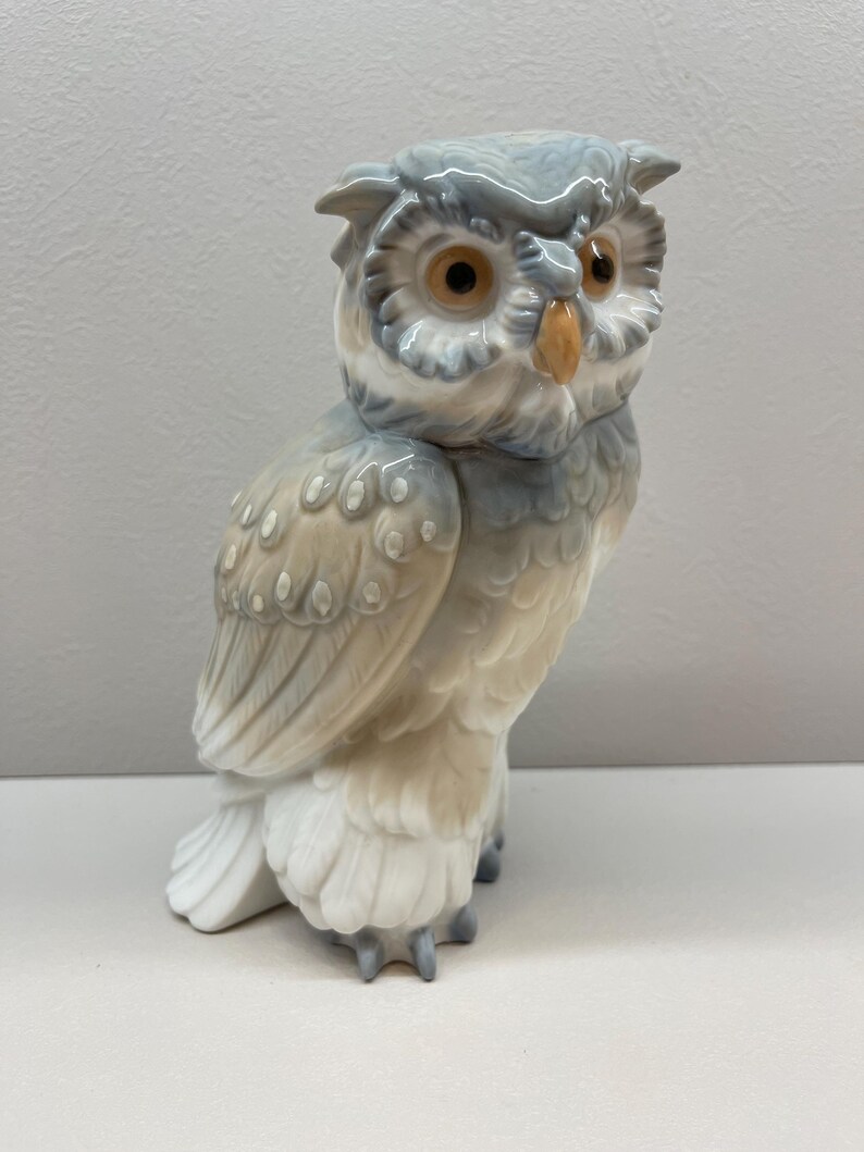 K&ouml;nnte beinhalten: Eine Eulenfigur aus Porzellan mit gl&auml;nzender Oberfl&auml;che in den Farben sanftes Grau, Wei&szlig; und Beige. Die Eule hat gro&szlig;e, runde, bernsteinfarbene Augen und einen kleinen, spitzen gelben Schnabel sowie detaillierte Federstrukturen am K&ouml;rper.