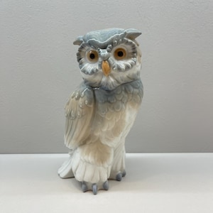 K&ouml;nnte beinhalten: Eine Eulenfigur aus Porzellan in einer sanften Farbpalette aus ged&auml;mpftem Grau, Creme und Wei&szlig;. Die Eule weist detaillierte Federstrukturen, markante orangefarbene Augen und einen gelben Schnabel auf und steht aufrecht mit leicht gedrehtem Kopf.
