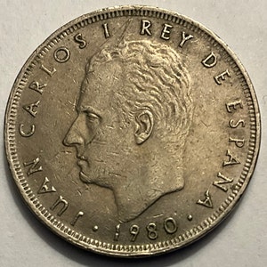 May include: Silver coin featuring a profile portrait of Juan Carlos I Rey de Espana, the King of Spain, with the year 1980 inscribed below.