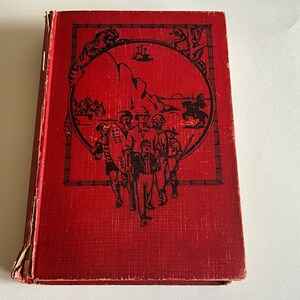 Op de afbeelding: Rood hardback boek met een zwart-witte illustratie van een groep mensen die voor een bergketen staan. De illustratie is omgeven door een zwart frame met een bamboe ontwerp. De titel van het boek is niet zichtbaar.