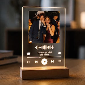 Peut inclure: Une plaque rectangulaire en acrylique transparent montée sur un socle en bois clair, présentant une photo illuminée d'un couple en tenue de soirée. Sous l'image, un graphique blanc affiche une onde sonore, le titre I'd rather go blind et Etta James.