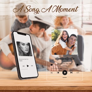 May include: A digital music player with a transparent glass screen displaying the song "Someone Like You" by Adele. The screen also shows a photo of a couple, a play button, and other music controls. The player is sitting on a wooden surface.