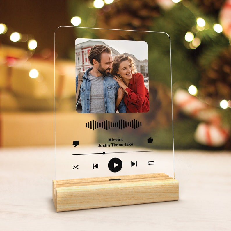 May include: A clear acrylic music player with a photo of a couple in the background. The music player has a black and white design with a play button, rewind button, fast forward button, and a volume control. The music player is on a wooden base. The song title is "Mirrors" by Justin Timberlake.