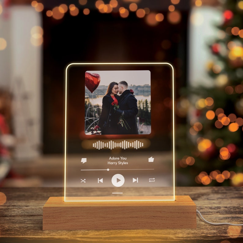 May include: A clear acrylic music player with a photo of a couple in front of a red heart balloon. The player has a play button, a volume control, and a song title that reads "Adore You Harry Styles".
