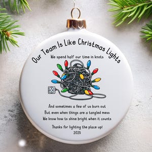Peut inclure: Boule de Noël blanche avec le texte "Our Team Is Like Christmas Lights". L'ornement présente une guirlande lumineuse emmêlée avec une prise. Le texte comprend l'année 2025.