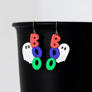 May include: Halloween-themed earrings with the word "BOO" spelled out in colorful letters. The letters are red, blue, and green, and are paired with white ghost-shaped cutouts. The earrings hang from silver hooks.