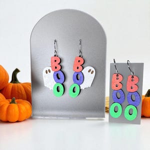 May include: Halloween-themed earrings featuring the word "BOO" in red, purple, and green, with a white ghost design. The earrings are displayed on a gray stand, with pumpkins in the background. The earrings are made of wood.