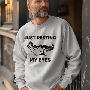 May include: A gray sweatshirt with the text "JUST RESTING MY EYES" and a graphic of a person reclining in a chair with their feet up.