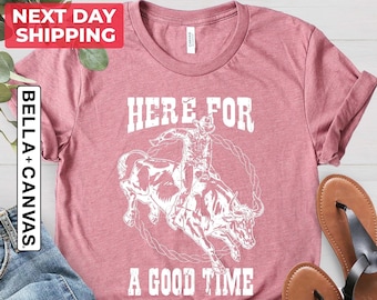 May include: Heather mauve t-shirt with a white graphic of a rodeo rider on a bull, encircled by a rope design. The text "HERE FOR A GOOD TIME" is printed below the graphic. The shirt is paired with blue jeans and brown sandals.