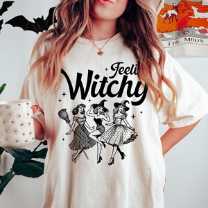 May include: An off-white t-shirt featuring a black graphic print of three women in witch hats and dresses with the text "Feelin' Witchy" above them. The design includes small star accents and a broomstick illustration, printed on a short-sleeved crew neck shirt.