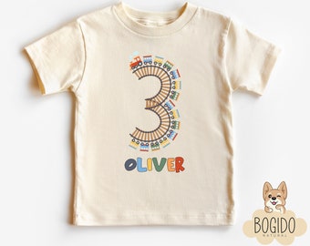 Puede incluir: Camiseta crema para niño pequeño con un diseño colorido de vías de tren que forma el número 3. El nombre "OLIVER" está impreso en colores arcoíris debajo. Un diseño divertido y lúdico para el cumpleaños de un niño.