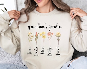 May include: Beige sweatshirt with the text "grandma's garden" and floral illustrations. Below the flowers are the names Jessica, Charles, Lilium, Katelyn, and Hannah. The sweatshirt is a neutral color.