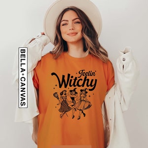 May include: A burnt orange t-shirt featuring the black text Feelin Witchy and a graphic illustration of three figures in witch hats and vintage-style dresses. The shirt includes a vertical BELLA+CANVAS brand label on the left side.