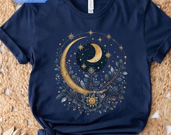 Può includere: Una t-shirt blu navy con una grafica centrale raffigurante una grande mezzaluna dorata e decorata, circondata da foglie blu scuro, un fiore in piena fioritura e stelle dorate. Il design include una mezzaluna più piccola su un cielo notturno.