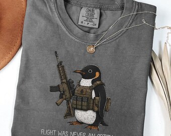 Può includere: Una t-shirt grigio antracite con la grafica di un pinguino che indossa un gilet tattico e impugna un fucile. Il testo sotto il pinguino recita "FLIGHT WAS NEVER AN OPTION". La t-shirt presenta un girocollo e un'etichetta Comfort Colors.