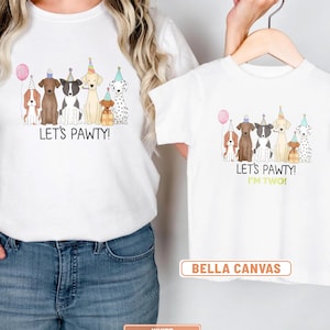 May include: White t-shirts with a dog-themed birthday party design. The adult shirt says "Let's Pawty!" and the child's shirt says "Let's Pawty! I'm Two!" The shirts feature illustrations of dogs wearing party hats and holding a pink balloon.