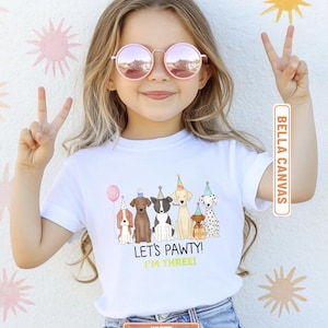 May include: White Bella Canvas t-shirt featuring a colorful illustration of six cartoon dogs wearing party hats and holding a balloon. The text "LET'S PAWTY! I'M THREE!" is printed below the dogs.
