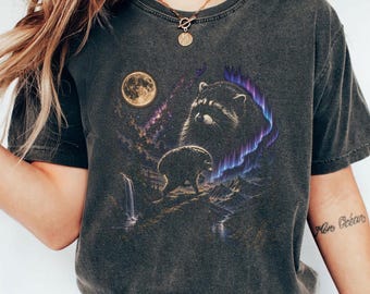 May include: A charcoal gray short-sleeve t-shirt featuring a graphic print of a raccoon, a full moon, a waterfall, and purple aurora borealis lights over a mountain landscape. The shirt has a crew neckline and a vintage-style washed finish.