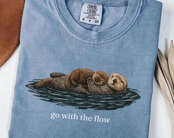 Può includere: Una t-shirt color blu jeans con una grafica di due lontre marine marroni che galleggiano nell'acqua. Il testo "go with the flow" è stampato in bianco sotto le lontre. La maglietta presenta un'etichetta del marchio Comfort Colors sul colletto.