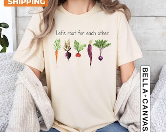 May include: A cream-colored short-sleeve t-shirt featuring a watercolor illustration of five different root vegetables in a row. Above the vegetables, the text reads Let's root for each other in black lettering. The shirt has a crew neckline.