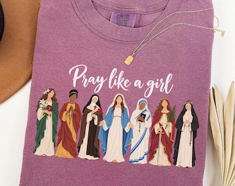 May include: A berry-colored t-shirt with a crew neckline featuring the text Pray like a girl and a graphic illustration of seven diverse religious figures in various robes and habits. The shirt has a Comfort Colors brand label at the inner neck.