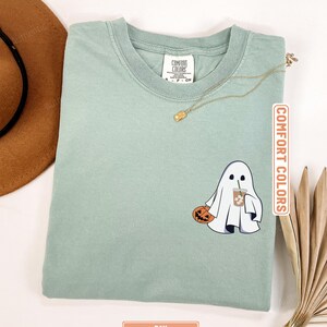 May include: A sage green t-shirt featuring a graphic of a white ghost holding a carved orange pumpkin and an iced coffee drink. The shirt has a crew neckline and a Comfort Colors brand label on the inside collar. The color is labeled as Bay.