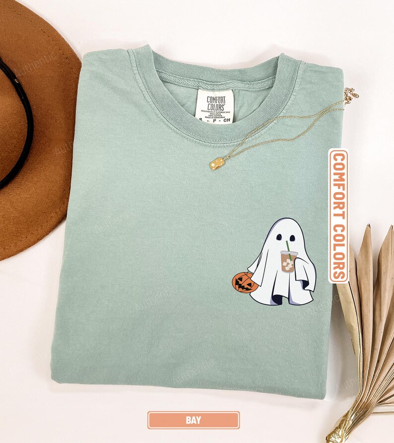 May include: A sage green t-shirt featuring a graphic of a white ghost holding a carved orange pumpkin and an iced coffee drink. The shirt has a crew neckline and a Comfort Colors brand label on the inside collar. The color is labeled as Bay.