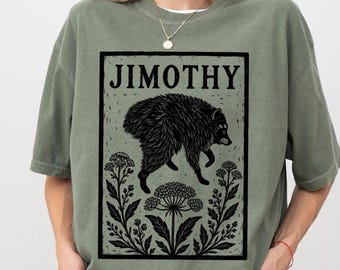 Può includere: Una t-shirt a girocollo color verde muschio con una stampa grafica nera di un procione sopra elementi floreali all'interno di una cornice rettangolare. Il testo JIMOTHY è stampato in un carattere serif in grassetto nella parte superiore.