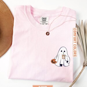 May include: A light pink t-shirt featuring a graphic of a white ghost holding a carved orange pumpkin and a clear plastic cup with a green straw. The shirt has a crew neckline and a Comfort Colors brand label printed on the inside neck area.
