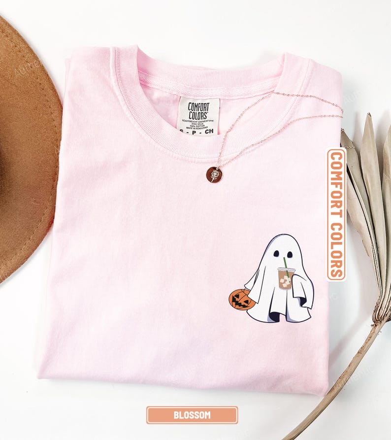 May include: A light pink t-shirt featuring a graphic of a white ghost holding a carved orange pumpkin and a clear plastic cup with a green straw. The shirt has a crew neckline and a Comfort Colors brand label printed on the inside neck area.