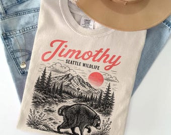 Può includere: Una t-shirt color avorio con una stampa grafica con il testo Timothy e Seattle Wildlife. Il design include un'illustrazione in bianco e nero di un procione in una foresta con montagne e un sole rosso. Sono visibili un cappello beige e jeans blu.