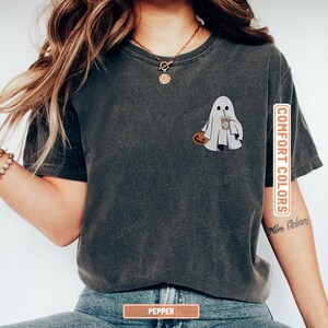 May include: A charcoal grey short-sleeve t-shirt featuring a graphic of a white ghost holding a small orange pumpkin and a brown iced coffee drink. The shirt has a crew neckline and a vintage-washed appearance with the text COMFORT COLORS.