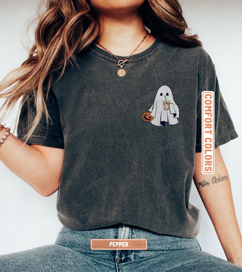 May include: A charcoal grey short-sleeve t-shirt featuring a graphic of a white ghost holding a small orange pumpkin and a brown iced coffee drink. The shirt has a crew neckline and a vintage-washed appearance with the text COMFORT COLORS.