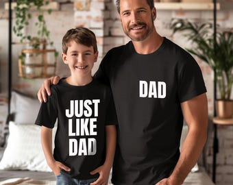 May include: Two black short-sleeved t-shirts. One shirt features the white text "JUST LIKE DAD" in a stacked block font. The other shirt features the white text "DAD" in a block font on the upper left chest area. Both shirts have crew necklines.