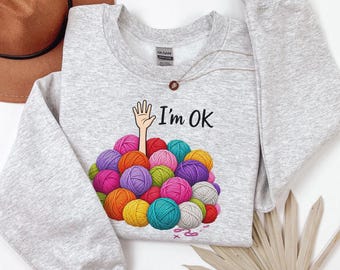 Può includere: Una felpa girocollo grigio chiaro con una grafica che mostra una mano che emerge da un mucchio di gomitoli di lana colorati con il testo I'm OK. Il design include gomitoli di lana in varie tonalità di rosa, blu, verde, giallo, viola e arancione.