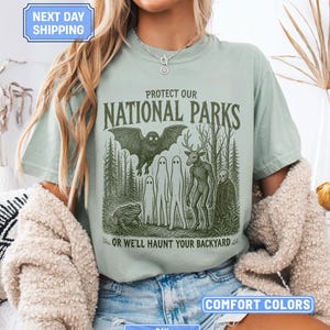 May include: A sage green t-shirt featuring a dark green graphic print of a forest scene with a bat, a toad, two ghosts, a deer-like creature, and a skeleton. The text reads PROTECT OUR NATIONAL PARKS OR WE'LL HAUNT YOUR BACKYARD in a dark green font.