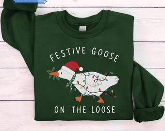 Può includere: Una felpa girocollo verde scuro con la grafica di un'oca bianca che indossa un cappello da Babbo Natale rosso e una sciarpa verde a motivi. L'oca è avvolta in luci natalizie colorate e tiene in bocca un rametto di agrifoglio. Testo: FESTIVE GOOSE ON THE LOOSE.