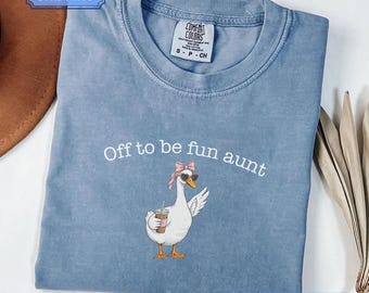 Può includere: Una t-shirt Comfort Colors color blu jeans con una grafica di un'oca bianca che indossa un foulard rosa e occhiali da sole neri mentre tiene in mano un caffè freddo. Il testo sopra l'oca recita, Off to be fun aunt. La maglia ha girocollo.
